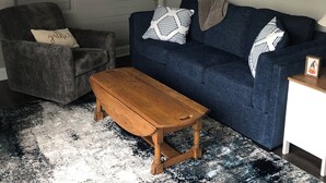 Ein Wohnzimmer mit einem dunkelblauen Sofa, einem Holztisch, einem grauen Sessel, einem weißen Beistelltisch, einer Stehlampe, einem Wandkompass und einem gemusterten Teppich.
