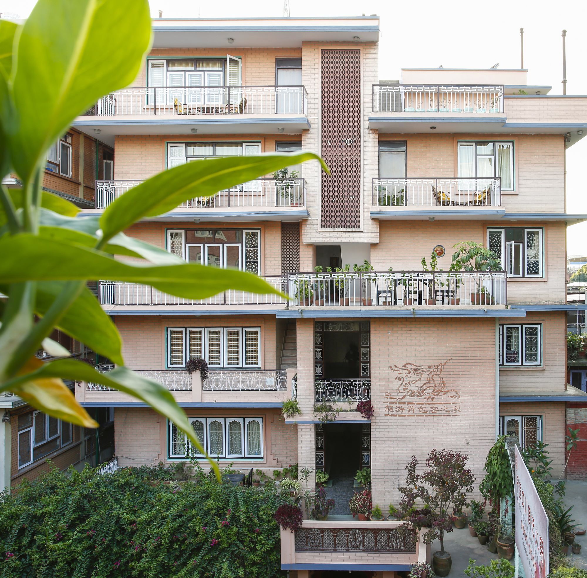 A multi-story building with balconies, a central vertical decorative element, and potted plants around the entrance.