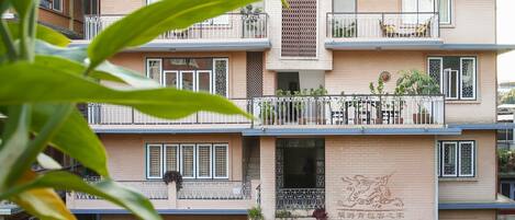A multi-story building with balconies, a central vertical decorative element, and potted plants around the entrance.
