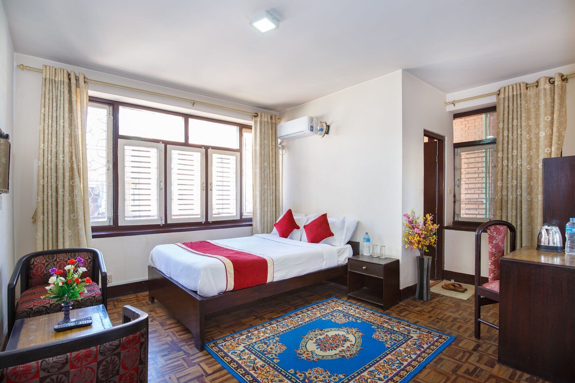 A hotel room with a bed, a desk, a chair, a small table with a vase of flowers, a water bottle, and a decorative rug.