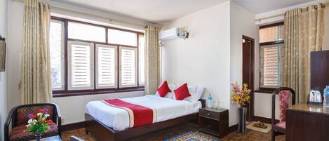 A hotel room with a bed, a desk, a chair, a small table with a vase of flowers, a water bottle, and a decorative rug.