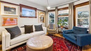A living room with a white sofa, a blue armchair, a round ottoman, and a red patterned rug. There are two windows with curtains, a potted plant, and framed artwork on the walls. - Gorgeous private home! Central Air 4 beds, hot tub, patio, designer furnishings (Portland)