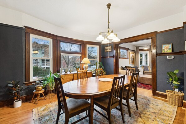 A dining area with a wooden table and chairs, surrounded by large windows and potted plants.