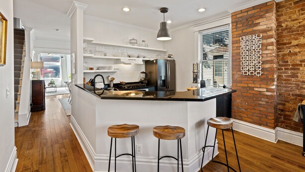 A modern kitchen with white cabinetry, a central island with stools, a brick accent wall, and a large window with blinds. - 
Upscale 3-Story German Village Home - Near Cafés (Columbus)