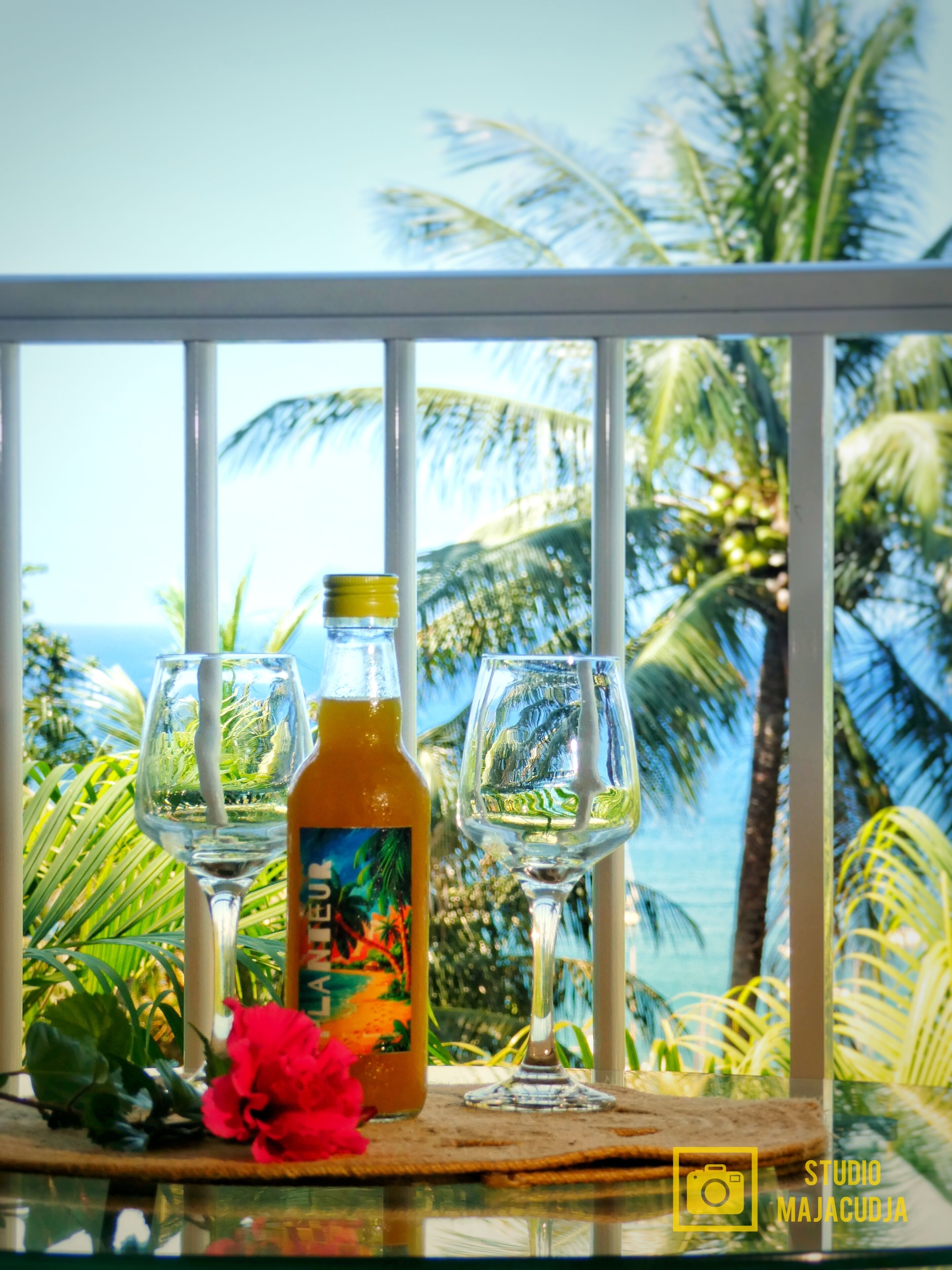 A table with two glasses and a bottle of drink, a red flower, and a view of palm trees and the ocean through a balcony railing.