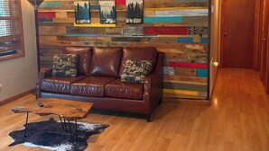 Un salon avec un canapé en cuir marron, une table basse en bois, un tapis à longues mèches et un mur en bois aux couleurs vives, orné de tableaux.