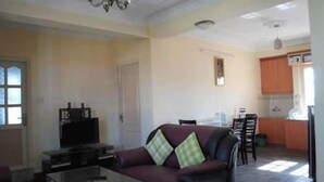 A living room with a sofa, a glass coffee table, and a television. There is a kitchen area with cabinets and a refrigerator. - Bliss Apartment (Kathmandu)