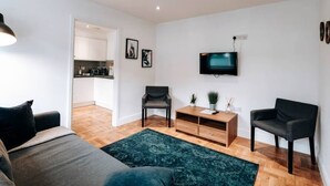 Una sala moderna con un sofá, dos sillones, una mesa de centro y un televisor. El suelo es de madera y hay una puerta que da a una cocina.