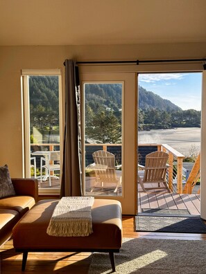 A modern living room with a sofa, a small table, and a view of a deck with two Adirondack chairs. - Ocean Cove Inn #7 - Ocean View (Yachats)