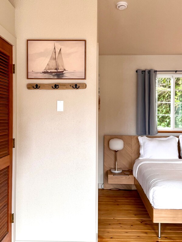 A bedroom with a wooden bed, a bedside table with a lamp, a window with curtains, a wall-mounted picture of a sailboat, and a wooden coat rack.