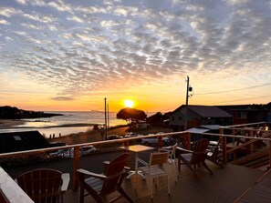 A wooden deck with chairs and tables overlooking a beach and ocean at sunset. - Ocean Cove Inn #6 - Ocean View (Yachats)