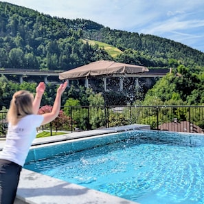 A swimming pool with a person in the foreground and a covered area with a view of a forested hillside in the background. - Hotel Schmuckhof (Chiusa)