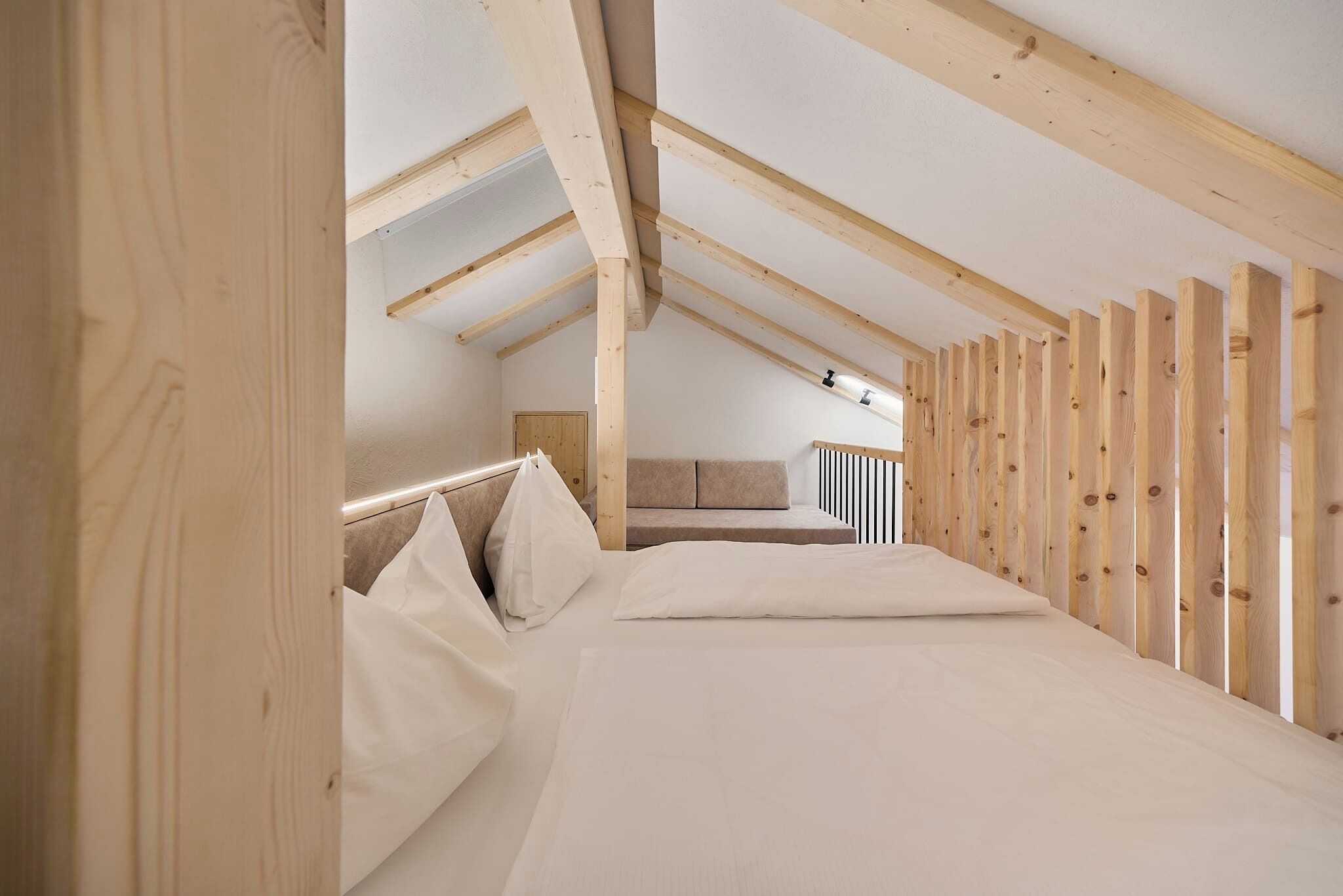 Ein Schlafzimmer mit schrägen Wänden, Holzbalken, einem Bett mit weißer Bettwäsche und einer Couch im Hintergrund.