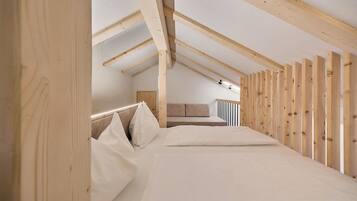 Ein Schlafzimmer mit schrägen Wänden, Holzbalken, einem Bett mit weißer Bettwäsche und einer Couch im Hintergrund.