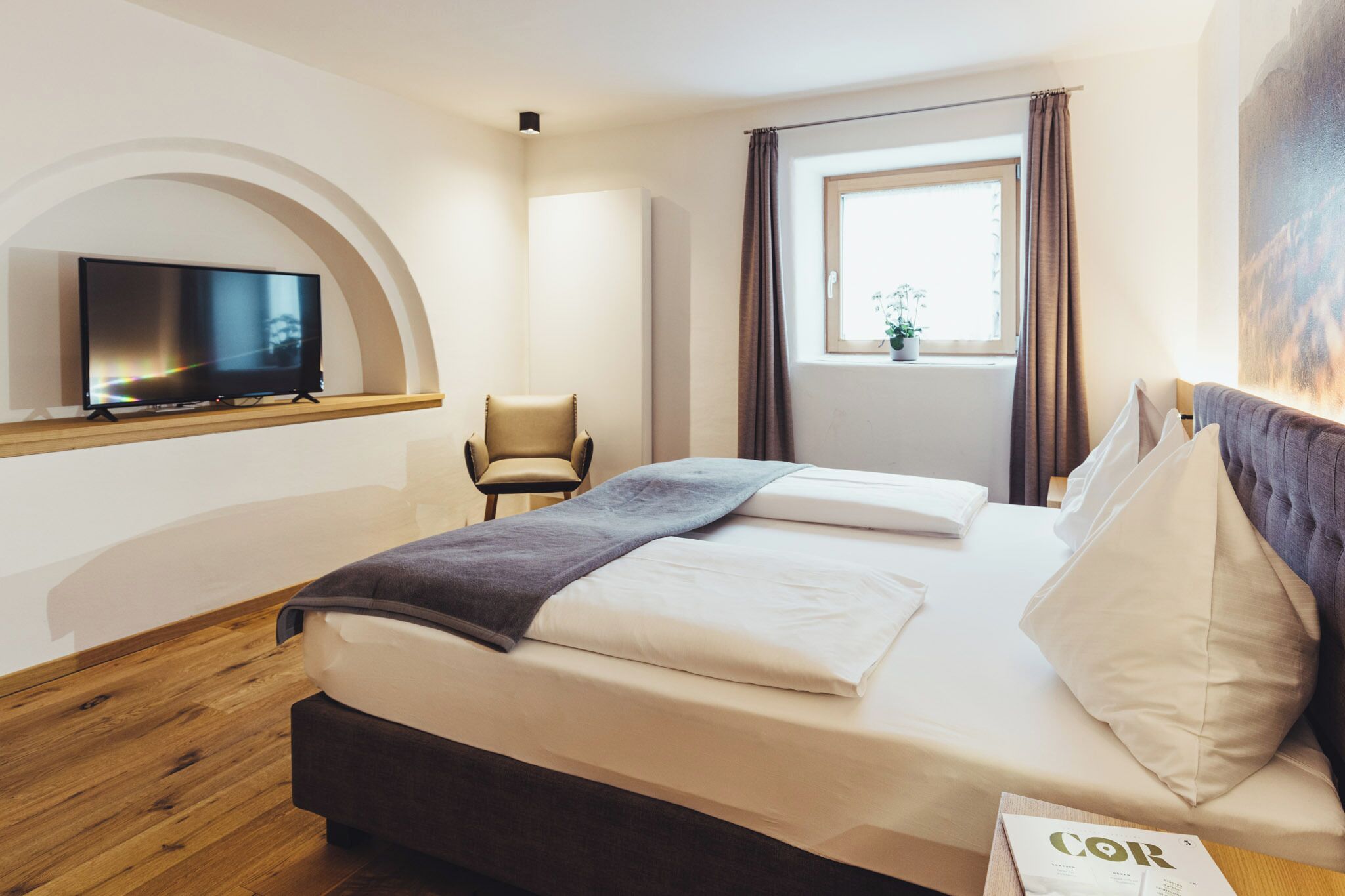 A hotel room with a bed, a television on a stand, a chair, a window with curtains, and a painting on the wall.