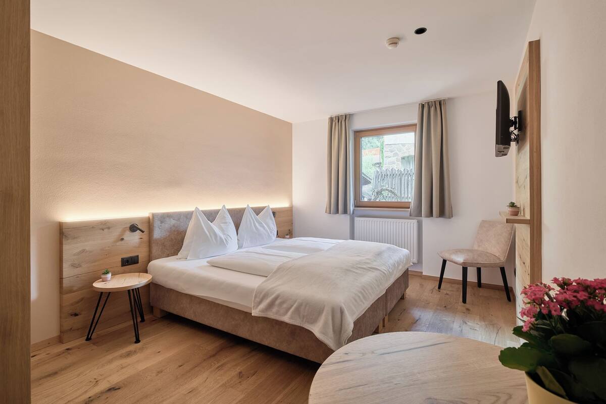 Une chambre moderne avec une tête de lit en bois, un lit avec une literie blanche, une table de chevet ronde, une chaise et une fenêtre avec des rideaux.