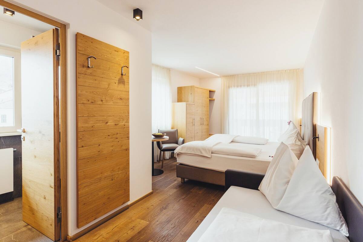 Une chambre d’hôtel avec une armoire en bois, un lit avec du linge de maison blanc, une petite table avec une chaise et une fenêtre avec des rideaux.