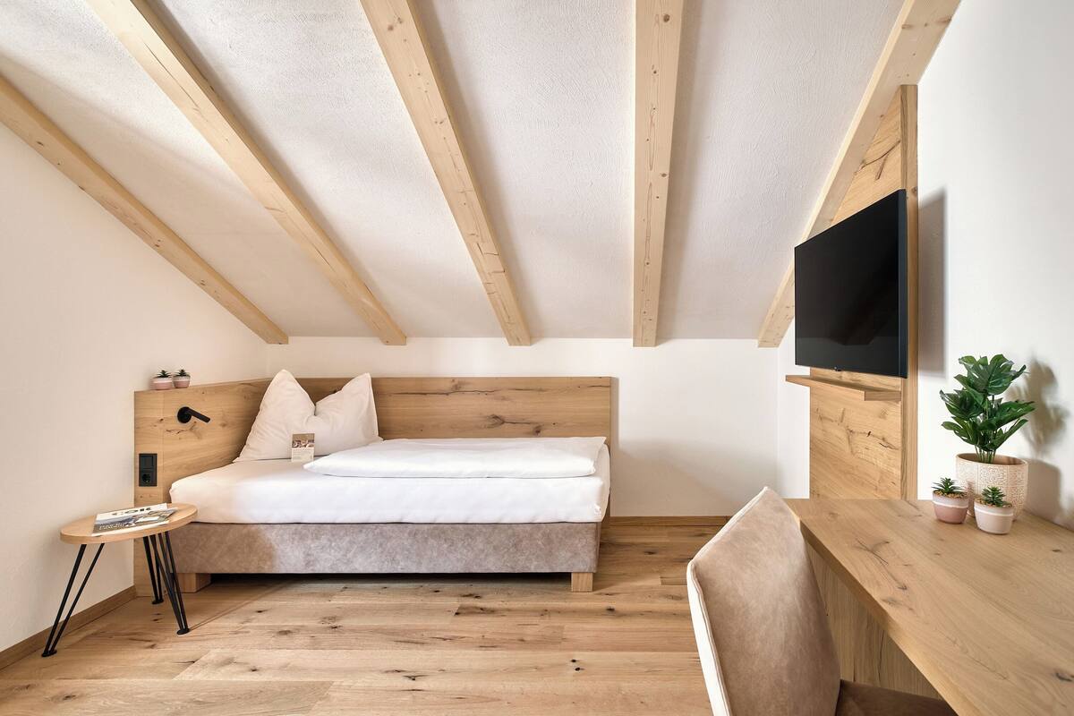 Une chambre avec un plafond en bois, un lit, une télévision, une petite table et un bureau agrémenté de plantes.