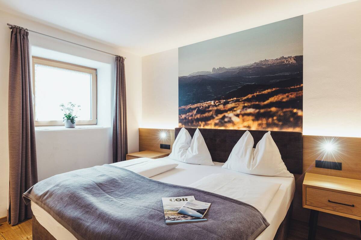 Une chambre d’hôtel avec un grand lit, une fenêtre avec des rideaux, une table de chevet avec une lampe et un tableau représentant un paysage accroché au mur.