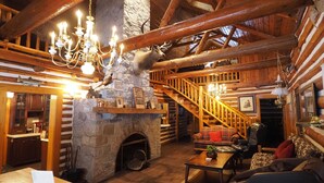 A rustic log cabin interior with a stone fireplace, wooden beams, a staircase, and a cozy seating area. - Secluded 10-Acre Riverfront Lodge. Pet-Friendly! Hot Tub! (Traverse City)