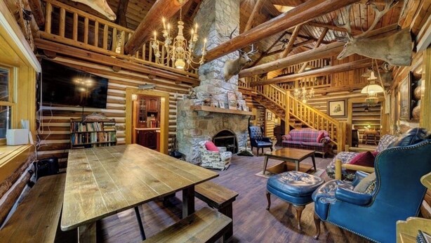 A rustic cabin interior with a stone fireplace, wooden beams, a dining table, and leather seating. - Secluded 10-Acre Riverfront Lodge. Pet-Friendly! Hot Tub! (Traverse City)