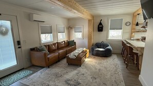 Ein modernes Wohnzimmer mit einem braunen Ledersofa, einem grauen Sitzsack und einer Essecke mit Holzbarhocker.