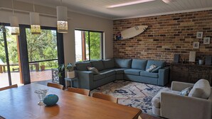 A modern living room with a large wooden table, a grey sectional sofa, and a brick wall with a surfboard. - Manyana Beach Getaway:
Spacious house with large deck 2 mins walk to the beach. (Manyana)