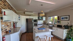 A modern kitchen with white cabinets, a stainless steel refrigerator, a microwave, and a gas stove. There is a wooden island with stools, a hanging light fixture, and a window with a view of greenery. - Manyana Beach Getaway:
Spacious house with large deck 2 mins walk to the beach. (Manyana)