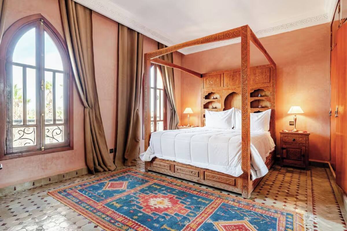 A four-poster bed with a patterned rug, a window with curtains, and a nightstand with a lamp.