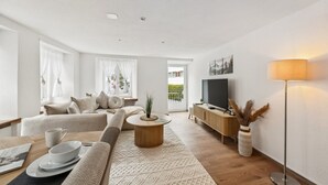 A modern living room with a sofa, coffee table, and TV. There is a window with curtains and a view of the outside.