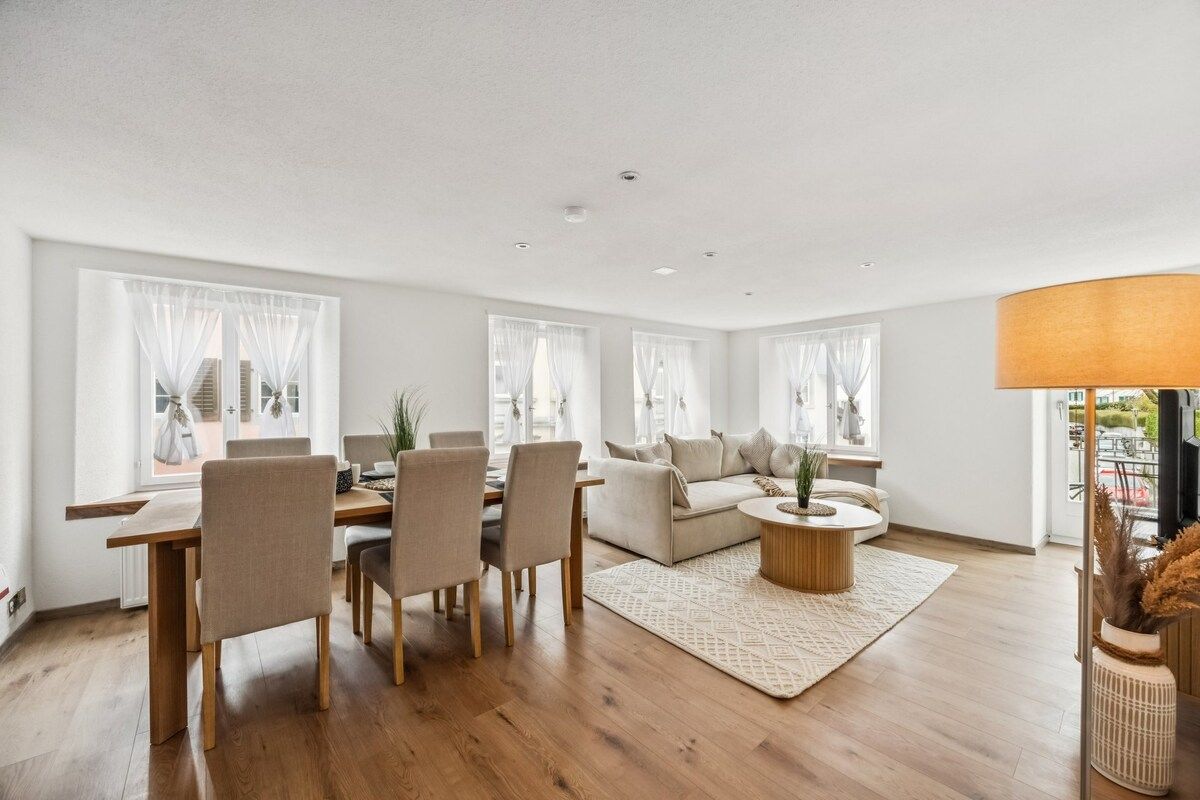 A modern living room with a dining area, large windows with sheer curtains, a round coffee table, and a sofa.