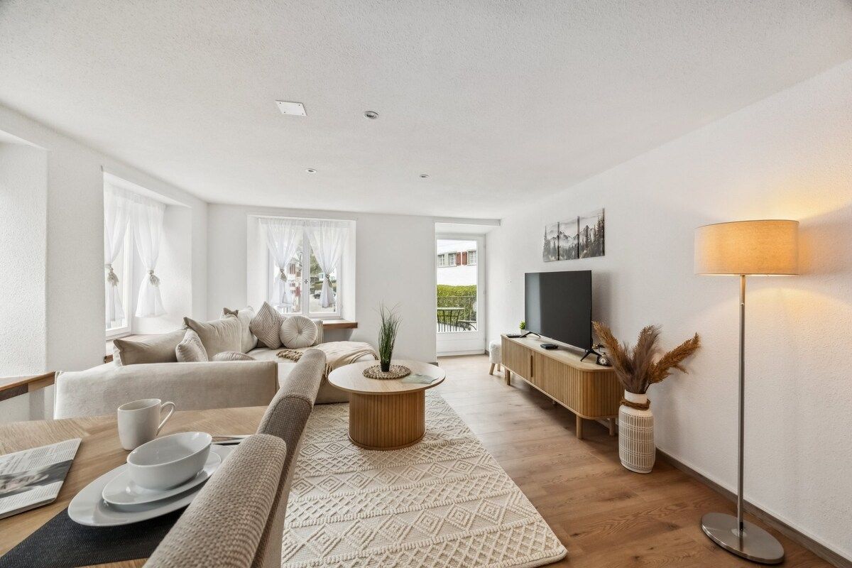 Una sala moderna con un sofá, una mesa de centro y un televisor. Hay un comedor con una mesa y sillas. La habitación tiene ventanas amplias con cortinas.