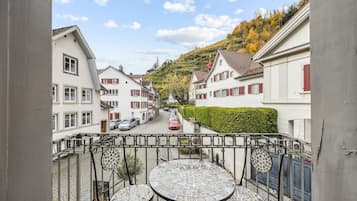 Ein Balkon mit einer gefliesten Tischplatte und Stühlen mit Blick auf eine Straße, auf der Autos geparkt sind und Häuser stehen.