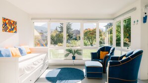 Un salón luminoso con un sofá blanco, sillones azules y un ventanal con vistas a árboles y edificios.