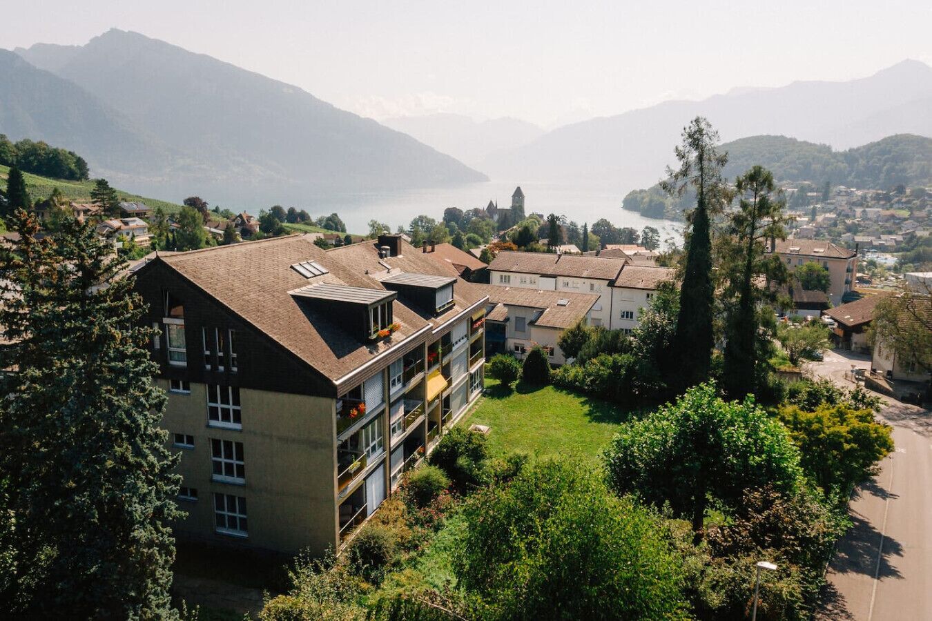 Un edificio de varios pisos con balcones, rodeado de vegetación y vistas a un lago y montañas.