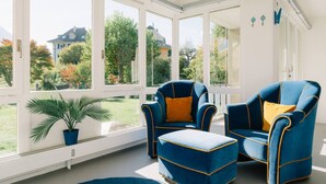 Una habitación iluminada por el sol, con ventanas amplias, dos sillones azules con detalles amarillos, un reposapiés a juego y una planta en maceta.
