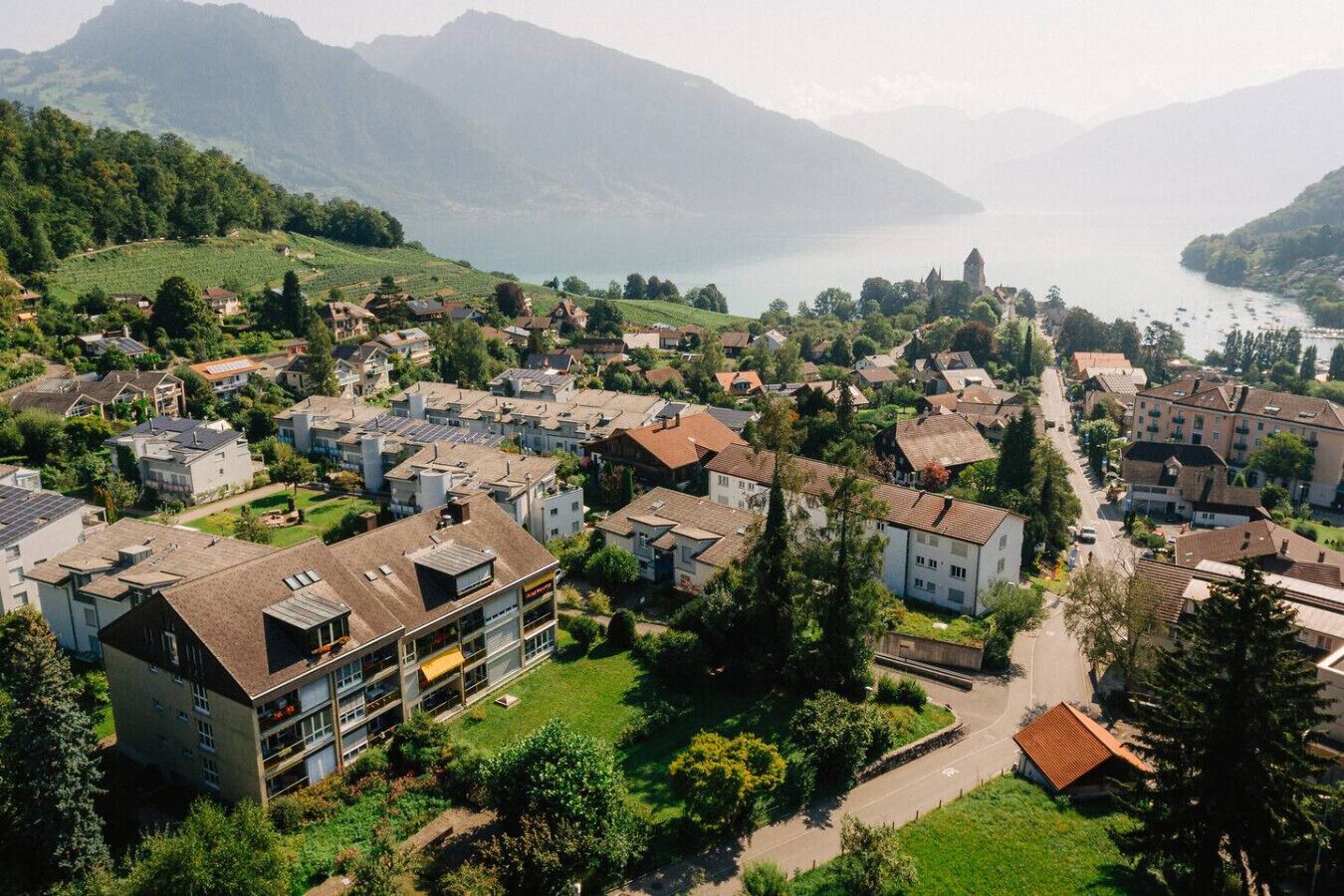Ein Wohngebiet mit Häusern und einem modernen Gebäude mit Balkonen, umgeben von Grünflächen und Bergen.