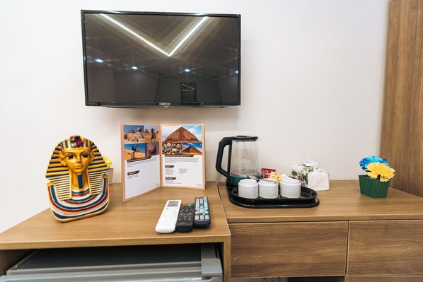 A hotel room with a flat-screen TV mounted on the wall, a remote control, a coffee maker, a tray with cups, a decorative Egyptian headpiece, and informational brochures. - Oriental Pyramids & GEM View (Giza)