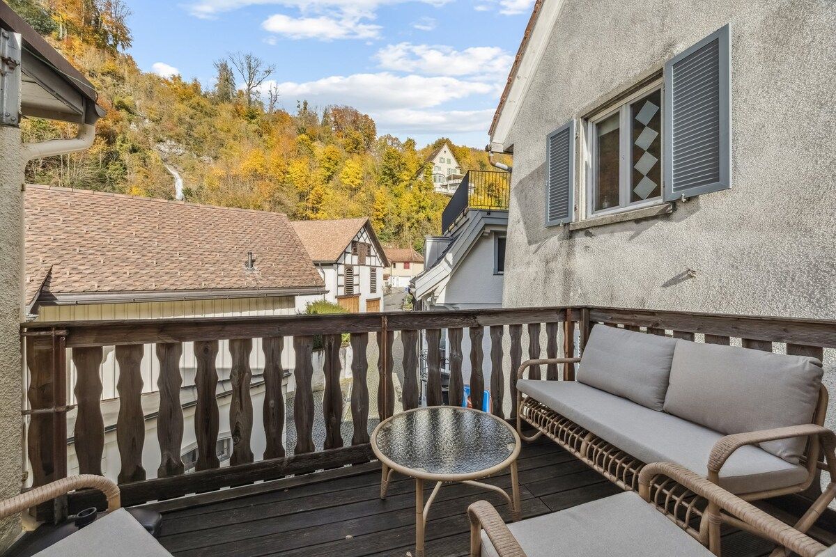 Un balcon avec un canapé en osier, une petite table ronde et une vue sur les maisons et les arbres.