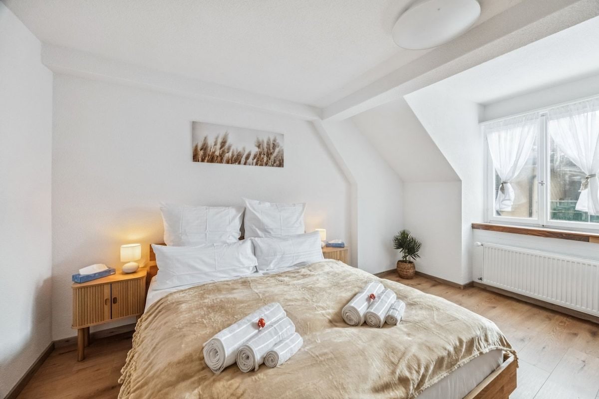 Un dormitorio con una cama grande, una mesita de noche con lámpara, una planta pequeña y una ventana con cortinas.