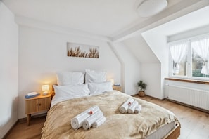 A bedroom with a large bed, a bedside table with a lamp, a small plant, and a window with curtains.