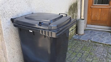 Uma lixeira preta com rodinhas, colocada sobre um calçamento de pedra ao lado de um prédio com porta de madeira e janela.