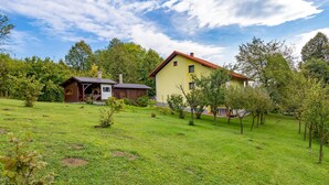 Una casa de dos pisos amb teulada vermella i façana groga, envoltada d'un jardí verd i arbres.