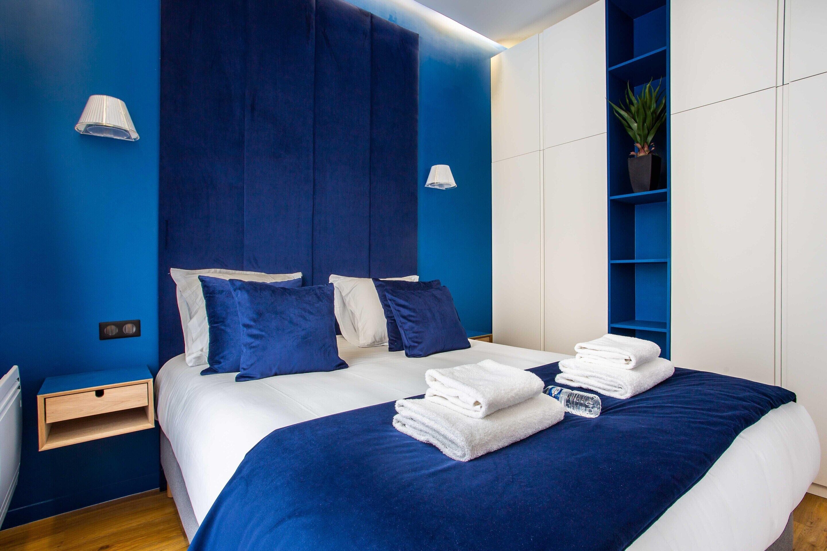 A modern hotel room with a large bed, blue and white bedding, a nightstand, a wall-mounted shelf with a plant, and a white wall lamp.