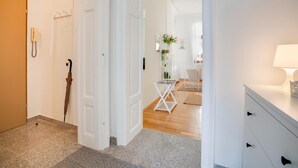 A hallway with a wooden door, a white wall-mounted phone, a black umbrella, a white folding chair, and a white desk with drawers. - Arcadio Home - Junior Suite (Graz)