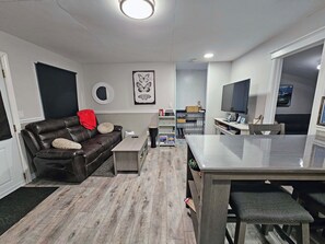 Ett modernt vardagsrum med en lädersoffa, ett matbord och en köksdel med en tv och en bokhylla.