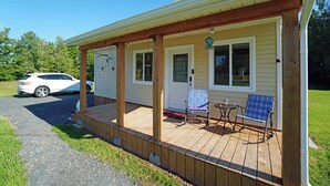A covered porch with two rocking chairs and a small table, a white car parked outside, and a house with a yellow exterior. - Tiny Home| Full Kitchen | Sleeps 4 + Work Desk. Relax, explore Acadian Peninsula (Allardville Parish)