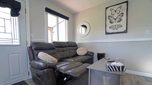 A living room with a reclining sofa, a small table, and a butterfly artwork on the wall. - Tiny Home| Full Kitchen | Sleeps 4 + Work Desk. Relax, explore Acadian Peninsula (Allardville Parish)