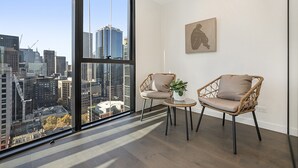 Una habitación con un ventanal amplio que ofrece vistas a la ciudad, dos sillones de mimbre, una mesita con una planta y una obra enmarcada en la pared.