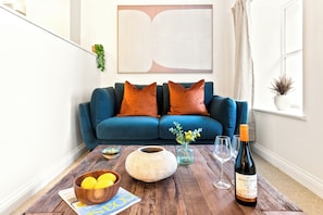 A modern living room with a blue sofa, orange cushions, a wooden coffee table, a vase with flowers, and a bottle of wine. - Boutique apartment with parking | Ye Old Mill (Penryn)